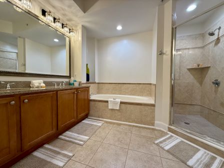 A bathroom with a double sink vanity, bathtub, and glass-enclosed shower. It has beige tiles, mirrors, and overhead lights.