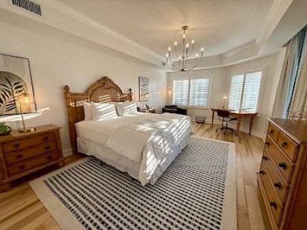 A well-lit bedroom with a bed, wooden furniture, a striped rug, and a chandelier, featuring windows with shutters and a cozy desk area.
