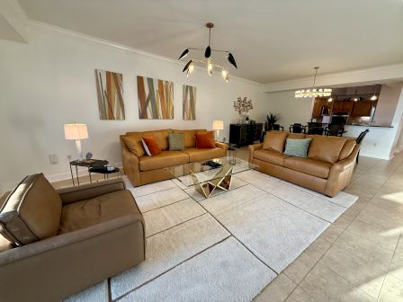 A modern living room with tan sofas, a glass coffee table, abstract wall art, and contemporary lighting. Kitchen visible in the background.