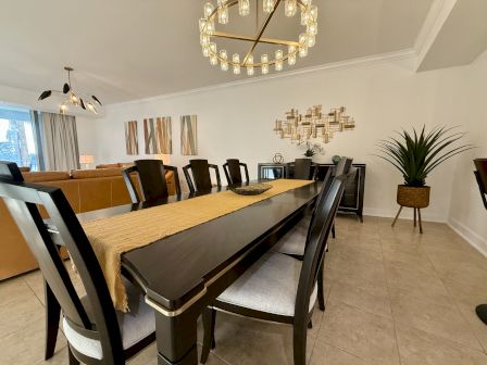 A modern dining room with a dark wooden table, a beige runner, and stylish chandeliers. Artwork and plants decorate the space.