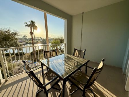 A sunlit patio with a glass table and chairs overlooks a scenic landscape featuring trees and a tranquil body of water.