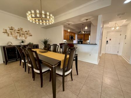 A dining room with an elegant chandelier, table for eight, decorative wall art, plants, and a view into the kitchen area.