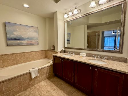 A bathroom with a large mirror, double sink vanity, bathtub, tiled walls, and a painting on the wall.