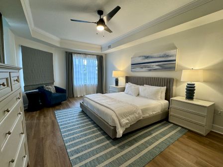 A modern bedroom with a bed, striped rug, two nightstands with lamps, a dresser, and a chair by the window with curtains.