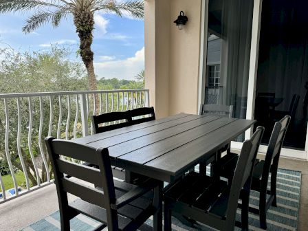 Outdoor patio with a black table and chairs, palm trees, and a view through the railing. A sliding glass door is nearby.