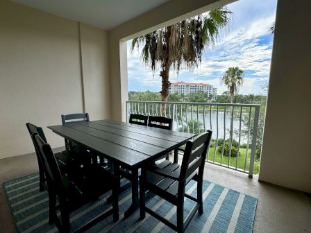 A balcony with a dining table and chairs overlooks a scenic view of greenery, palm trees, water, and a building in the distance.