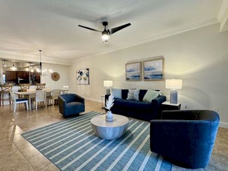 A modern living room with blue chairs, striped rug, art, and dining area with a chandelier. Neutral tone decor.