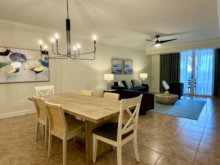 A modern living-dining area with wooden dining table, abstract art, dark sofas, a chandelier, and a ceiling fan near large windows.