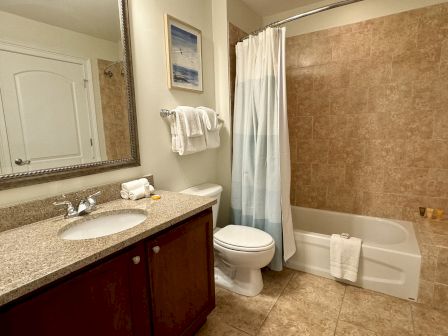 A clean bathroom with a granite countertop, sink, mirror, toilet, and a bathtub with a shower curtain and tiled walls.