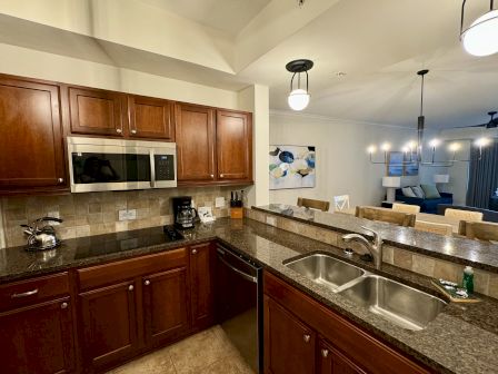 A modern kitchen with wooden cabinets, granite countertops, a microwave, a kettle, and a double sink. Dining area visible beyond.