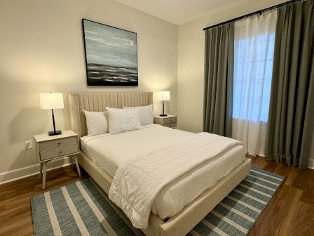 A cozy bedroom with a bed, bedside tables with lamps, a painting, and curtains over a window, featuring a striped rug on the floor.