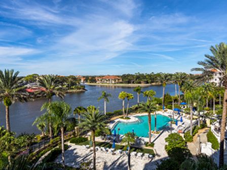 A scenic view of a resort with a pool, surrounded by palm trees, and a lake with houses in the background under a clear blue sky.