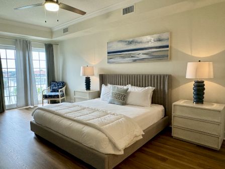 A modern bedroom with a large bed, bedside tables with lamps, a ceiling fan, an armchair, and a window with curtains.