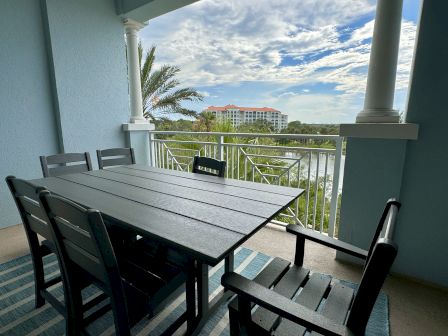 Outdoor patio with a black dining table and chairs on a balcony. Scenic view of palm trees and a building in the background.