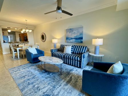 A cozy living room with blue furniture, striped sofa, coffee table, and dining area under a chandelier. Kitchen visible in the background.