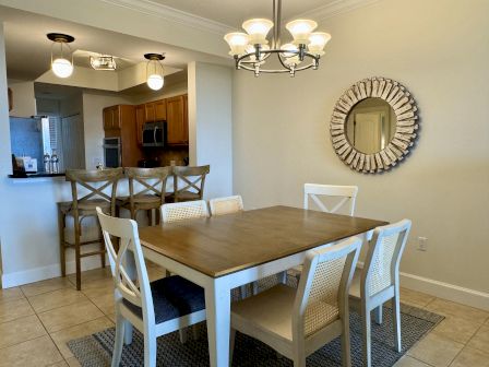 A dining area with a wooden table, six chairs, a decorative mirror, and a kitchen bar with stools. Bright lighting overhead.
