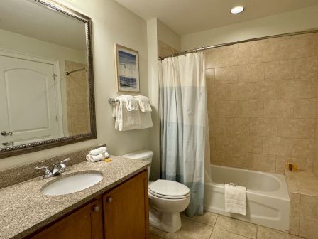 A clean bathroom with a granite countertop, sink, mirror, toilet, and shower with curtain; towels and toiletries are neatly arranged.