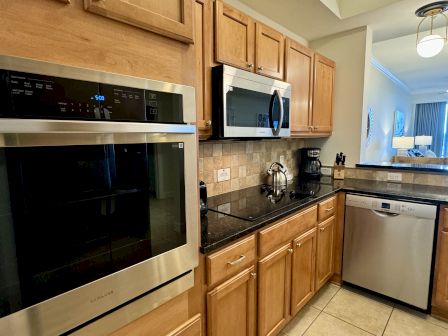 A modern kitchen with stainless steel appliances, wooden cabinets, a black countertop, a kettle, and a coffee maker on a tiled backsplash.