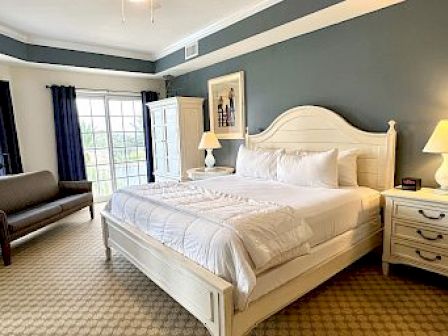 A bedroom with a bed, nightstands with lamps, a couch, a wardrobe, and large windows with curtains is shown in the image.