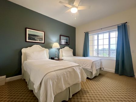 A bedroom with two twin beds, white bedding, bedside lamp, framed art, ceiling fan, and a window with blue curtains and a patterned carpet.