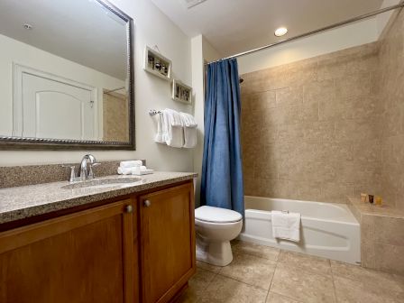 A bathroom with a large mirror, countertop, sink, toilet, blue shower curtain, and tiled bathtub area with towels arranged neatly.