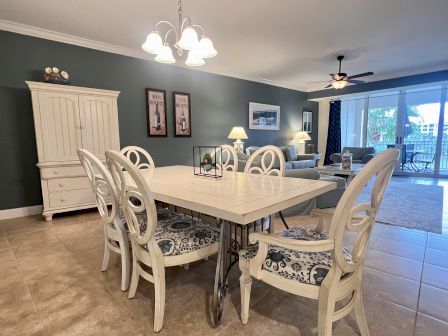 A dining room with a table and chairs, a cabinet, wall art, and a living area with a sofa, lamps, and balcony doors.