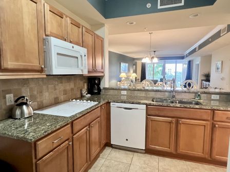 A kitchen with wooden cabinets, white appliances, granite countertops, and a view into a dining area with a large window.