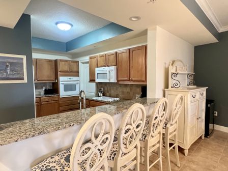 A kitchen with granite countertops, wooden cabinets, a microwave, oven, sink, chairs, and a small storage unit in a cozy setting.
