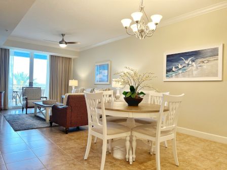 A cozy living room and dining area with a ceiling fan, artwork, potted plant, and elegant chandelier near a large window leading to a balcony.