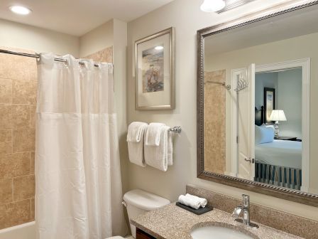 A bathroom with beige tiles, a shower curtain, towels, a mirror reflecting a bed, and wall art above the towels.