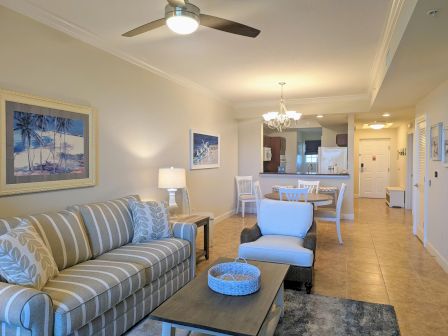 A cozy living room with a striped sofa, armchair, coffee table, dining set, ceiling fan, wall art, and kitchen in the background.