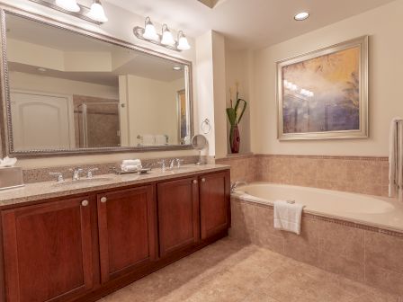 A bathroom with a large mirror, double sink, bathtub, art piece above tub, towel, and decorative vase on counter under soft lighting.