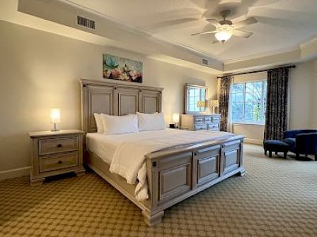 A spacious bedroom features a large bed, two nightstands, lamps, and a window with curtains. Neutral tones and a ceiling fan complete the look.