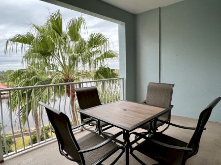 A balcony with a table and four chairs overlooks a lush palm tree and a serene water view.