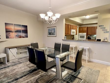 A modern dining area with a glass table, six chairs, chandelier, barstools, and kitchen. Wall art showcases a sunset and beach scene.