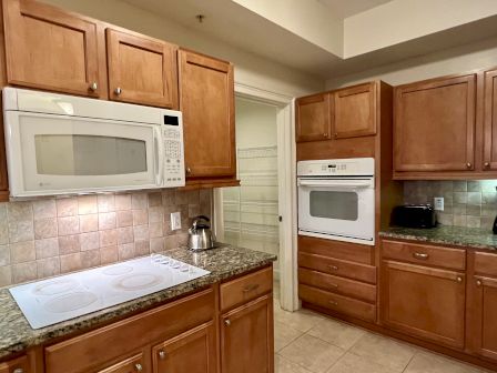 This image shows a kitchen with wooden cabinets, a white microwave, oven, stovetop, kettle, and toaster on granite countertops.