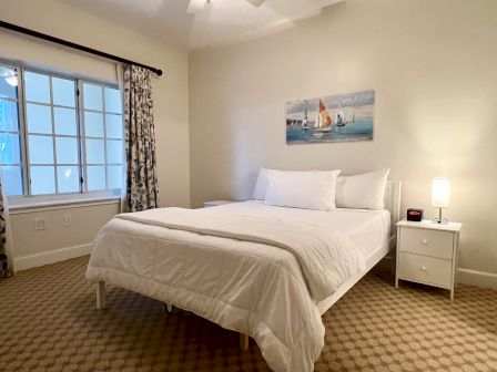 A cozy bedroom with a bed, white linens, nightstand, lamp, clock, window, curtains, and a painting of sailboats on the wall.