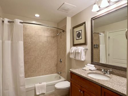 The image shows a clean, modern bathroom with a bathtub, shower curtain, towels, a sink with a granite countertop, and a large mirror.