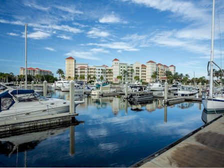 A marina with boats docked, calm waters reflecting the sky, and buildings in the background under a partly cloudy blue sky.