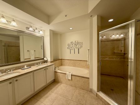 A spacious bathroom features a large mirror, double sinks, tub, and glass shower, with neutral tones and decorative wall art.