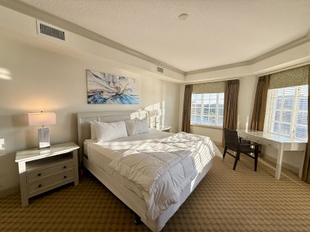 A bright bedroom with a large bed, nightstand with a lamp, desk with a chair, and windows with curtains, decorated with wall art.