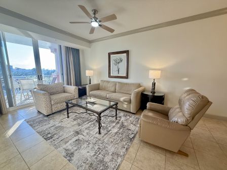A living room with a beige couch, armchair, glass coffee table, ceiling fan, and balcony view. Framed art and lamps add decor.