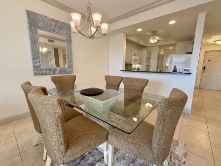 The image shows a dining area with a glass table, six brown chairs, a chandelier, a large mirror, and a view into a kitchen.