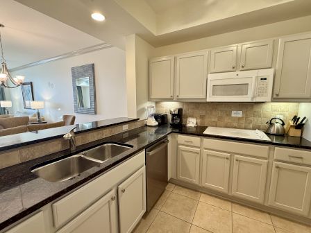 A modern kitchen with white cabinets, black countertops, a double sink, a microwave, and various appliances. Adjacent dining room visible.