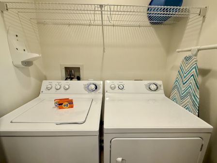 A laundry room with a washing machine, dryer, detergent box, ironing board, and a storage shelf above, all in a compact space.