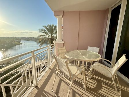 A sunlit balcony with a glass table, four chairs, and a view of a river, palm tree, and a bridge in the distance.