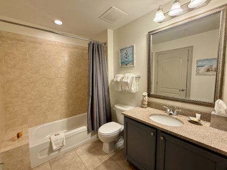 A bathroom with a tub, gray curtain, toilet, towels, and a granite sink counter. There are two wall art pieces and a large mirror.
