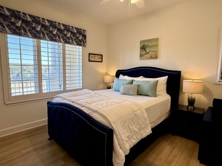 A cozy bedroom with a navy bed, white bedding, sea-themed decor, and two lamps by windows showcasing a scenic outdoor view.