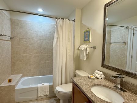 A bathroom with a shower-tub combo, beige tiles, a white shower curtain, towels, a large mirror, and a granite countertop sink.