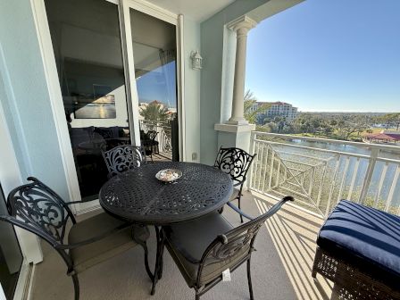 A balcony features a metal table with four chairs, seashell centerpiece, blue cushioned bench, overlooking a scenic view of buildings and water.